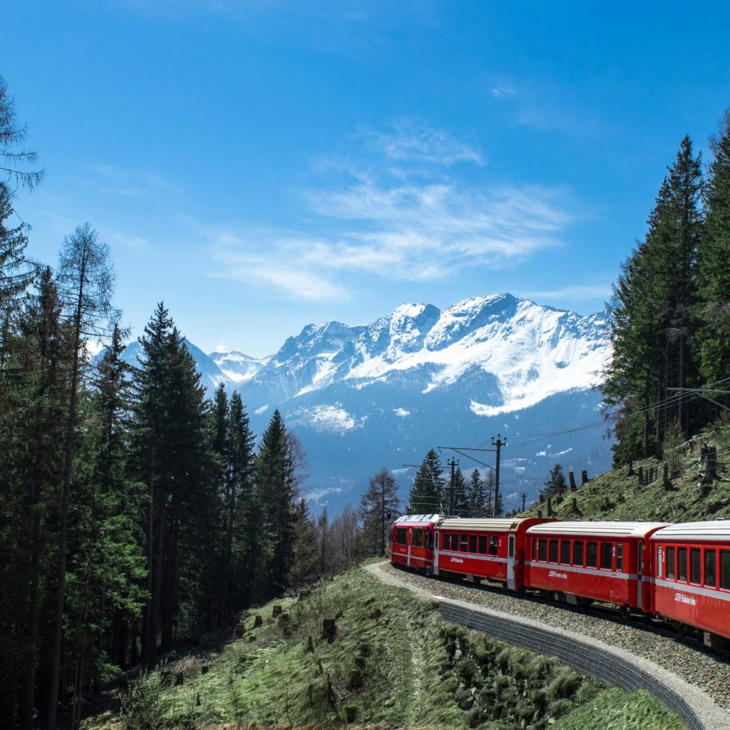 A red train travels through lush forests and snowy mountains under a clear blue sky in Switzerland.