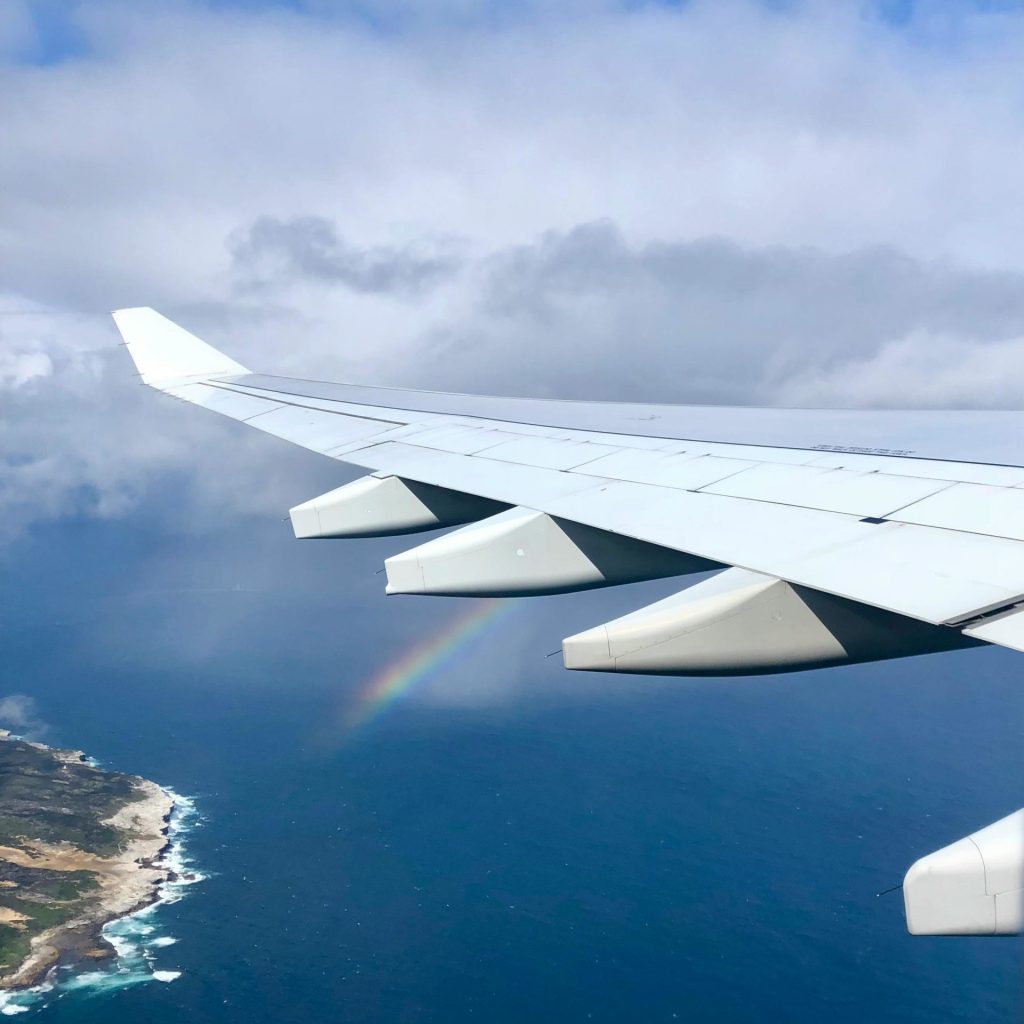 A scenic aerial view of an airplane wing over the ocean, featuring a rainbow in the sky.